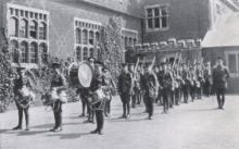 30 boys in military uniform, several holding drums, stand in lines. A uniformed gentleman watches over them.