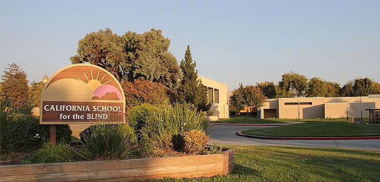 Photo of the main entrance to the California School for the Blind from Walnut Avenue, facing the school. The sign at the main entrance is shown with the theater and several other buildings in the background.