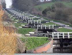 A series of approximately 20 black lock gates with white ends to the paddle arms and wooden railings, each slightly higher than the one below. On the right is a path and on both sides grass and vegetation.