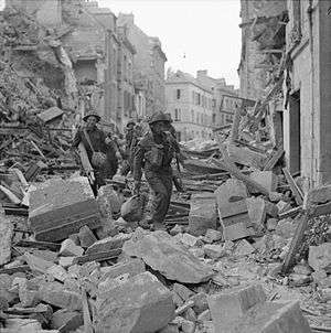 A line of soldiers clambering over the heaped rubble of destroyed buildings in a badly-damaged street.