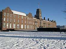 A photograph of the front of a large, wide, three-storey building made from light brown bricks. There are approximately thirty windows on each floor, all equally spaced out. There is a clock tower in the centre of the front of the building, which has a flagpole and flag on the top.