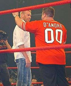 Two adult males standing in a red roped wrestling ring with a camera man standing in the foreground. One male is wearing a white shirt and bluejeans, with the other wearing a red shirt with the word "D'Amore" on the back and black pants.