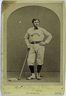A man, wearing a baseball uniform with the words "BOSTON" in the center, leans on a bat held in his right hand.