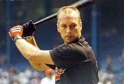 A man with short hair prepares to swing a baseball bat. He is wearing a black shirt with "Orioles" written in orange (obscured), and the bat is held over his right shoulder. He is wearing orange and black batting gloves on his hands.