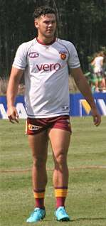 A black-haired man in a white shirt and maroon shorts stands on a football field.