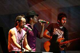 Three people. The first wearing a tank top, pink with a black legend at center, playing a keyboard and singing into a microphone with stand. The second person is wearing a maroon shirt and a gray hat with white stripes, while his left hand holds a microphone. The third person holding a guitar in his hand and wearing a black shirt with an ilustacion colored in white.
