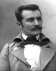 Monochrome head and shoulders photographic portrait of a man with a mustache and short dark hair, face turned slightly to his left, wearing a jacket, high-collared shirt, and loose bow tie