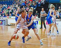A female basketball player is attempting to drive to a basket while another female player is guarding her, and attempting to reach for the ball.