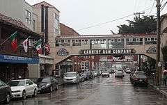 Tourist shops and historical remnants of the sardine canning industry line both sides of Cannery Row, which is busy even though it is raining. Two bridges that are labeled with the company names of defunct sardine canneries stretch over the street, connecting buildings on either side.