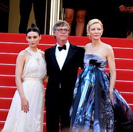 A man wearing a black tuxedo, white shirt, black bow tie, and eyeglasses is standing with his arms around the backs of two women. Behind them are red-carpeted steps. The woman on his right, a brunette, is wearing a haute couture white halter gown with ruffles and embroidery on the skirt; while the woman on his left, a blonde, is wearing a strapless ballgown with prints and shades of blue, light gray, black, and some hints of red.