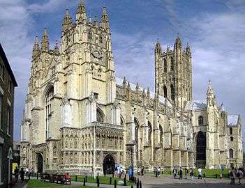 View of the exterior of Canterbury Cathedral on a bright day. The building is of pale stone with three large towers and much ornate Gothic detail. People are entering through a richly sculptured side porch.