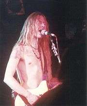 A male singer and guitarist, Jerry Cantrell, pictured onstage at a concert. He has an electric guitar strapped over his shoulder and he is singing into a microphone. He has long hair and he is not wearing any shirt.