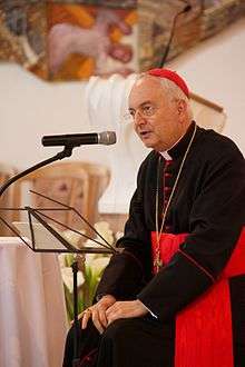 Man in robes of a Cardinal sitting before a microphone