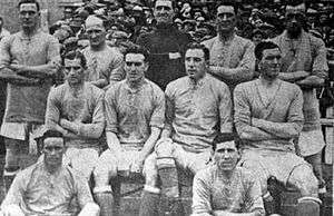 A football team of eleven men pose for a photograph. Five men stand with arms folded behind four men sitting on a bench. Two players sit in front of the bench on the floor.