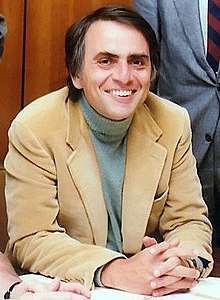 A middle-aged man sits at a desk, wearing a casual jacket and jumper. He is smiling and holding his hands together to pose for a photograph; this is physicist Carl Sagan.