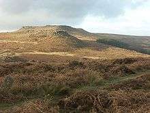 Photograph showing a moorland view. The moor is covered in heather of varying shades of brown. Stones are scattered across the moor. In the middle distance there is a rock outcrop atop a small hill. Behind it is a larger hill with a flat top.