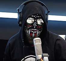 A painted man, wearing a black hoodie, sunglasses and headphones. He speaks into a microphone placed in front of him.