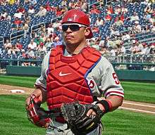 An olive-skinned man wearing a gray baseball jersey, red chest protector, red backwards baseball helmet, and sunglasses carrying a catcher's mask in his right hand and wearing a catcher's mitt on the left