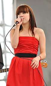 A young woman in long red dress performing live, while handling the microphone in her hand.
