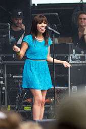 A Caucasian female with shoulder-length, wavy brown hair and wearing a powder blue dress sings and pearls into a microphone on stage, extending both arms in an emphatic gesture.