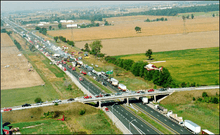 "Ontario Highway 401 viewed from high above travels into the distance from the bottom-right to the top-left. The Manning Road overpass crosses the freeway near the bottom of the image. The surroundings are entirely agricultural. On the highway, several dozen vehicles are piled into each other. The middle of the large pileup is smoking."