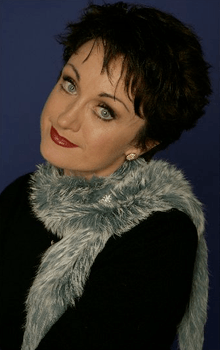 A close-up posed upper body shot of a woman. Her head is tilted to her left. She has short, black hair and pale blue eyes. She wears a grey/white fleecy boa/scarf over her black top. She has make up and red lipstick and sports an earring in her left ear (right ear is not visible).