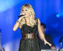 A blonde-haired young woman in a black dress singing into a microphone