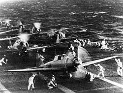 Planes on the deck of an aircraft carrier, with technical crews in white overalls attending the planes