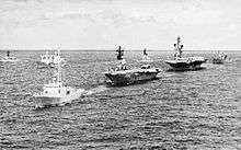 Warships sailing in a line on the open ocean. A small warship leads the line, followed by two aircraft carriers and a replenishment vessel. Other warships sailing in the same direction are in the background.