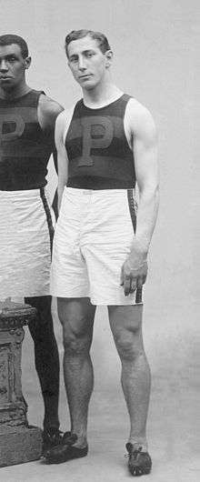 A white male stands and faces the camera in an older photograph. His right hand is behind his back. He is wearing a stripped sleeveless athletic top with a P on the front, white athletic shorts, and running shoes.