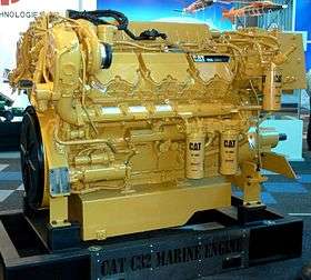 Caterpillar-brand engine encased in its yellow enclosure depicted. On the right there is a cylindrical oil filter and on slightly below the middle left also two oil filters side by side. "CAT 1R-1808 - Advanced" reads on these filters. On the bottom it reads "CAT C32 MARINE ENGINE". On the upper right there is a black plate reading "CAT C32" along with some unreadable text. Several white quality check stickers glued on the engine.