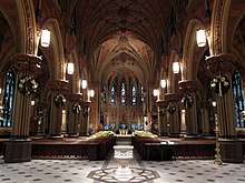 A church interior with a tall vaulted ceilings, wooden pews on a stone floor, and stained glass windows