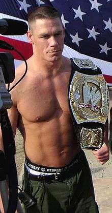 A man with a championship belt on his shoulder, with the American flag in the background.