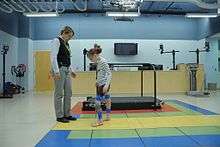 A girl wearing leg braces walks towards a woman.  They are standing in a gym, and a treadmill is visible in the background.