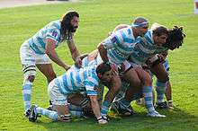 Facing right a group of seven men, in blue and white hooped jersey, bind together and crouch to form a scrum, the eighth player stands behind them observing the off-picture opposition.