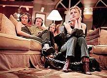 An image of four women and a man sitting on a couch in a living room. They are all chained together by the ankles.