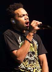 An image of a man holding a microphone close to his mouth and wearing a golden skull t-shirt.