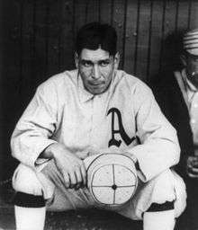 A dark-haired man in a white baseball jersey with a blackletter A on the chest sitting on a bench holding a white cap in his hands