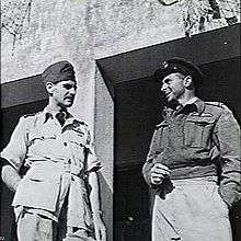Two men in military uniform standing outside of a building talking to each other. The man on the left is wearing a lighter coloured uniform with short sleeves. The man on the right is wearing a long sleeved, dark jacket.