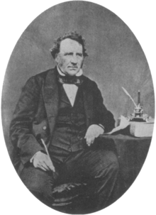 A middle aged Victorian gentleman sits beside a table wearing a dark suit with waistcoat and bow tie. His left elbow rests on the table. In his right hand he holds a quill pen.