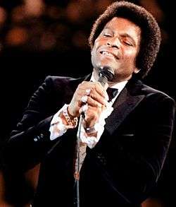 A dark-skinned man in a tuxedo singing into a microphone