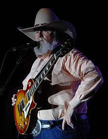 A grey-haired man with a large beard wearing a cowboy hat, holding a guitar and talking into a microphone