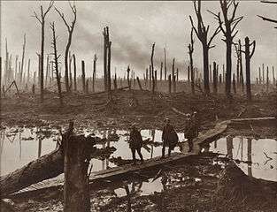A group of soldiers walk across wooden duckboards that have been constructed over a waterlogged and muddy field. Shattered trees dot the landscape, with a low lying haze in the background.
