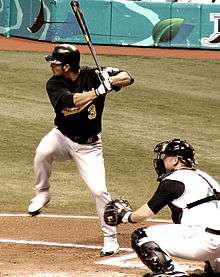 A man in a dark baseball jersey, batting helmet, and white pants takes a left-handed baseball swing while another man kneels wearing catcher's gear behind him.
