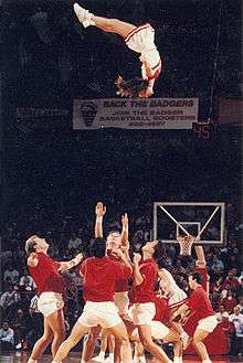 On a basketball court with a net and crowd in the background, four men in red shirts and white shorts look up, ready to catch a woman who is upside down about 3.5 metres (12 feet) in the air above them. A similar group can be partially seen behind them.