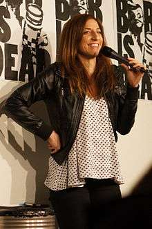 A smiling Chelsea Peretti, hand on hip and speaking into a microphone