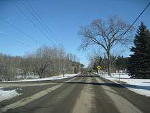 Ground-level winter view of a two-lane rural road heading straight into the background. A second two-lane road begins at this road and leaves to the left foreground.