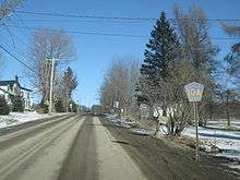 Ground-level winter view of a two-lane road ascending a hill. A blue and yellow route marker reading "Chenango County 10A" is on the right side of the road in the foreground, and a house is on the left side in the background.