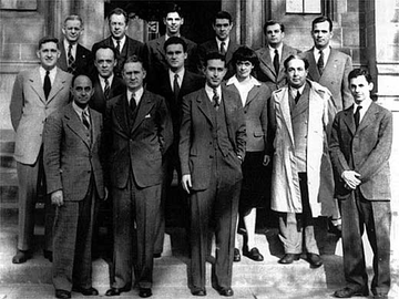 14 men and one woman, all wearing formal suit jackets, with Szilard also wearing a lab coat