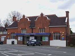 A brown-bricked building with a black roof and a blue sign that reads "CHIGWELL STATION" in white letters all under a white sky
